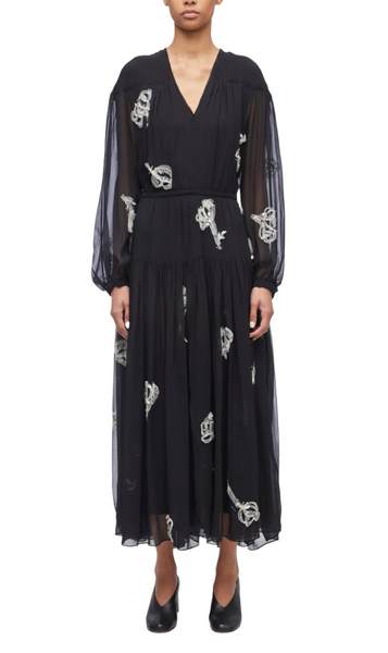 A person wearing a 3.1 Phillip Lim Deconstructed Floral Dress, characterized by its black long-sleeved V-neck design with white floral embroidery, standing against a plain background.