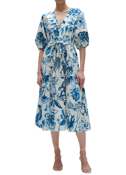 Woman wearing the Figue Joyce Dress, a white and blue floral wrap dress from Figue, featuring balloon sleeves, a cinched waist, and a V-neckline, paired with strappy heels.