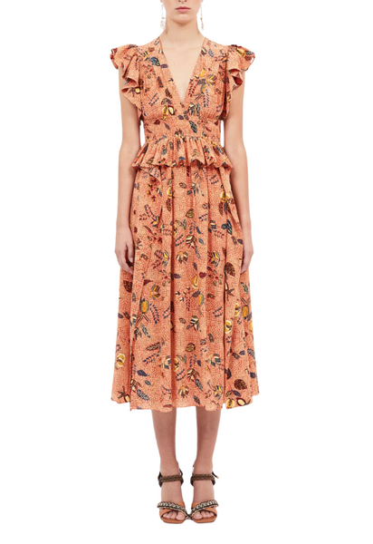 A person is wearing the Ulla Johnson Brielle Dress, a sleeveless, peach-colored dress adorned with ruffle details and a batik Coral Dahlia print by Ulla Johnson, standing against a plain white background.