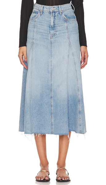 A person wearing the Mother The Full Swing Denim Skirt in light blue, knee-length, made from 100% cotton with a frayed hem, paired with a black ribbed sweater, standing against a plain white background.