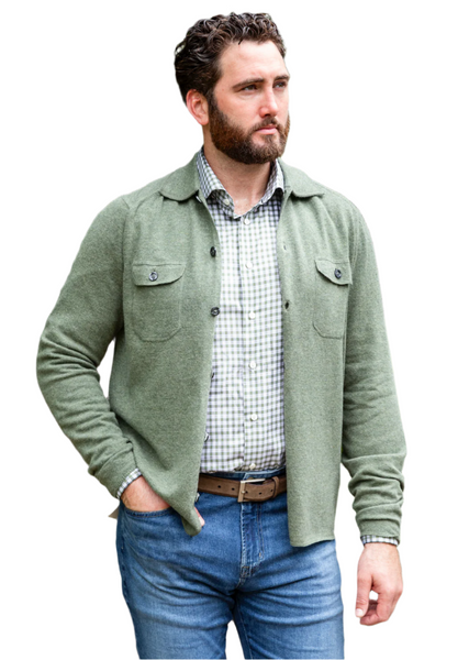 A bearded man wearing the Alan Paine Tore Button Knit Shirt in a green hue stands with one hand in his jeans pocket, the buttoned chest pockets adding a touch of rugged style.