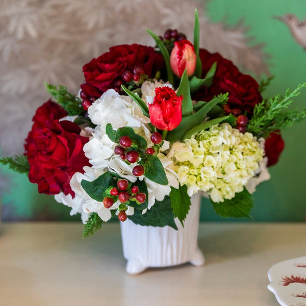 The Live Holiday Floral Arrangement by Hive Floral Studio showcases a stunning centerpiece of red roses, red tulips, white hydrangeas, green leaves, and berries beautifully arranged in a white vase on the table.