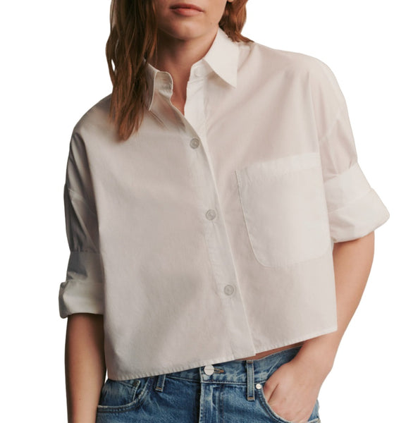 A person wearing a white, short-sleeved TWP Next Ex Shirt crafted from Italian cotton shirting with a front pocket and blue jeans. The shirt sleeves are rolled up.