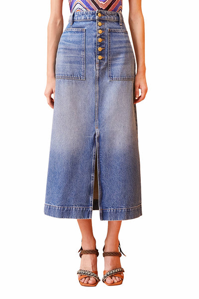 A person is wearing a long, high-waisted Ulla Johnson Bea Skirt made of non-stretch denim, featuring front buttons, a front split, and large front pockets. It's paired with a colorful top and brown sandals.