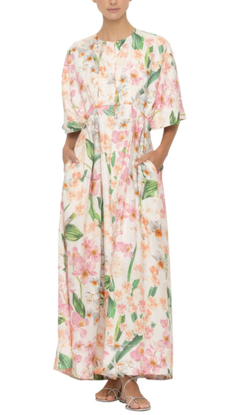 A person wearing a long, floral dress with short sleeves and sandals, standing against a plain white background showcases one of the coveted pieces: the Agua by Aguabendita Gesto Maxi Kaftan.