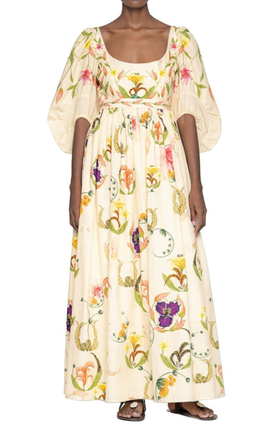 A person wearing the Agua by Aguabendita Vivianne Maxi Dress, a long, cream-colored floral print dress with puffy sleeves and colorful flower patterns, paired with black sandals.