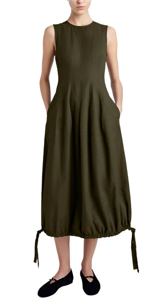 A woman stands wearing a sleeveless, olive green Proenza Schouler Marley Dress made of Tech Cotton. The ankle-length dress features handy side pockets and an adjustable bubble hem, paired with black flat shoes. Her hands are in the pockets.