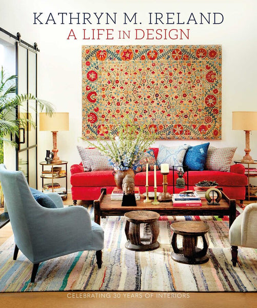 Cover of *"Kathryn Ireland: A Life in Design"* by Simon & Schuster, featuring a stylish living room with a red sofa, assorted chairs, a wooden coffee table, and a patterned wall tapestry by the renowned interior designer. Text reads "Celebrating 30 Years of Interiors.