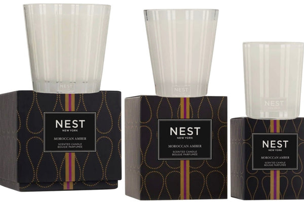 Image of two Nest NEST Moroccan Amber Candle Collections in glass holders, placed on their respective black boxes with a luxurious gold and purple pattern. The exquisite blend of amber, patchouli, and hints of bergamot creates a captivating scent.