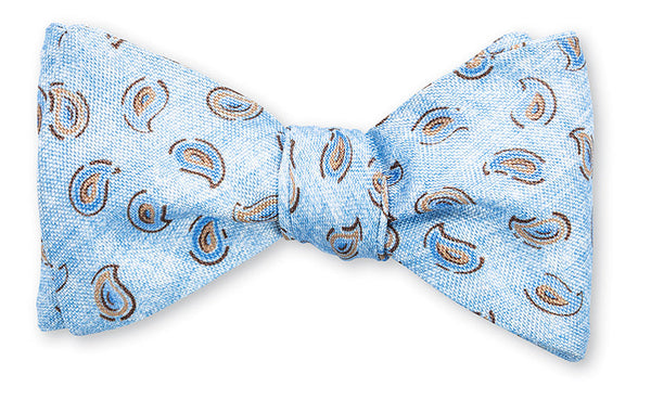 A hand-made light blue silk R. Hanauer Brinkley Pine bow tie with a pattern of brown and beige paisley designs on a white background.