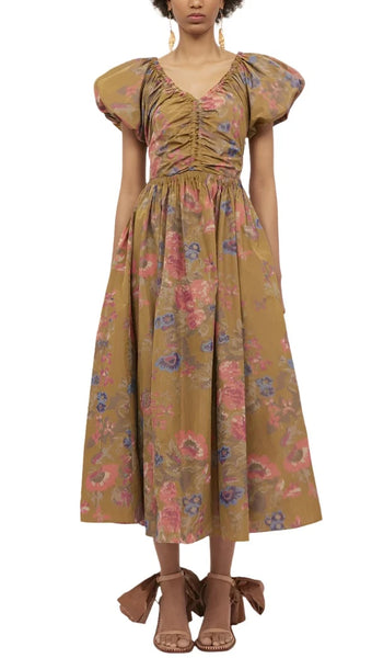 A person wearing an Ulla Johnson Cecile Dress, crafted from floral printed taffeta featuring puff sleeves and a ruched bodice with an elasticated waist, paired with brown bow-accented open-toe sandals.