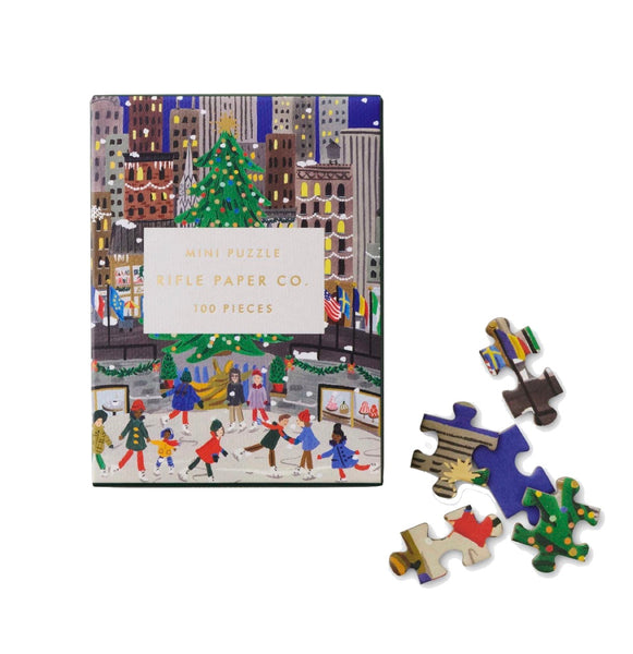 The Rifle Paper Co. Holiday On Ice Mini Puzzle features a delightful cityscape with people ice skating around a beautifully adorned tree, packed in an attractive tuck box. Five intricately crafted pieces, made from textured paper, lie next to the box.