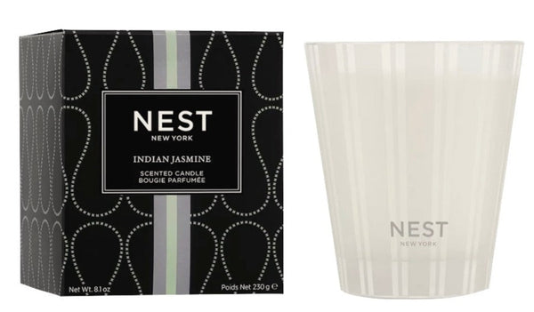 A white jasmine candle in a clear glass with "Nest" written on it, next to a black box labeled "NEST Indian Jasmine Classic Candle" by Nest. The premium wax box has a white and light green stripe design.