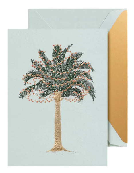 The "Crane Twinkling Palms Card Set" from Crane includes a card featuring an illustration of a palm tree decorated with string lights. An envelope is subtly visible behind, adding to the festive allure of this beach glass blue card.