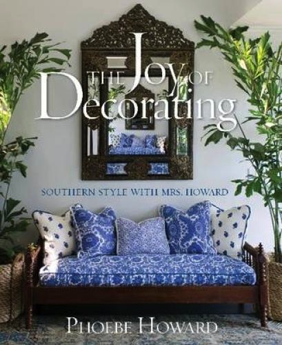 Book cover of "Joy of Decorating by Phoebe Howard" by Common Ground, featuring a luxurious and exclusive haven with a decorative mirror, blue and white patterned sofa, and vibrant green plants.