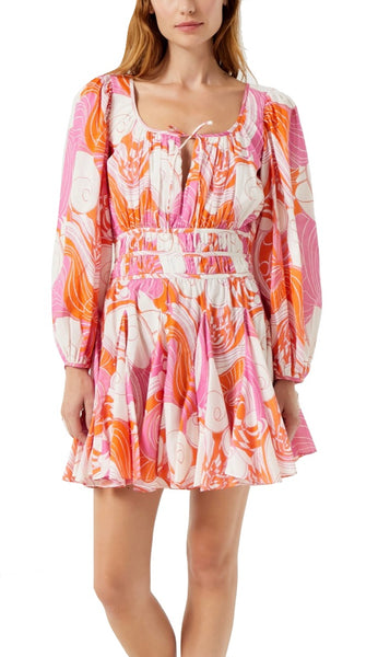 A woman is wearing a Rhode Leona Dress in a vibrant Pink Deco Surf print. Crafted from Cotton Cambric Fabrication, this colorful dress features pink, orange, and white floral patterns with long sleeves and a short, pleated skirt.