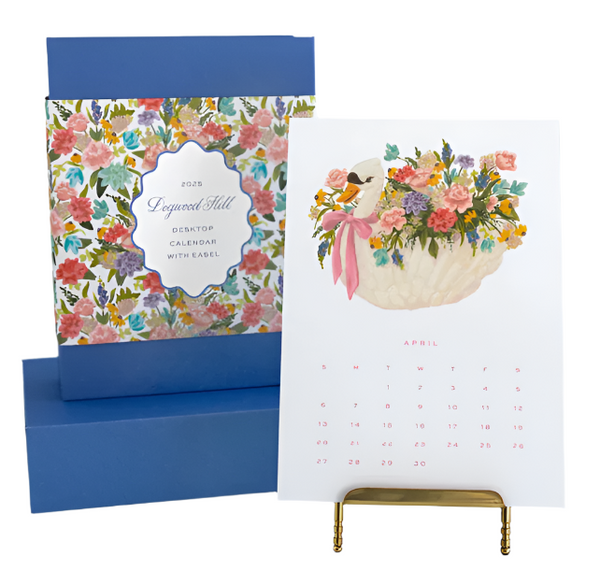 Presenting the Dogwood-Hill 2025 Desktop Calendar with a brass easel stand. Displayed is the April page, featuring original artwork of a bird and flower basket illustration. In the background, you can see the calendar box embellished with a floral design.