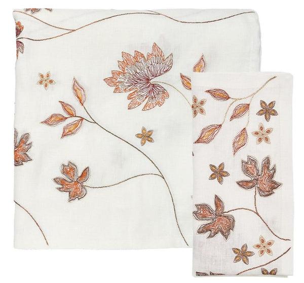 The Kim Seybert Maple Hues Collection tablecloth features a white fabric adorned with orange and brown floral embroidery, making it an ideal choice for a fall table setting. A large flower along with smaller blossoms are beautifully scattered across this elegant embroidered piece.