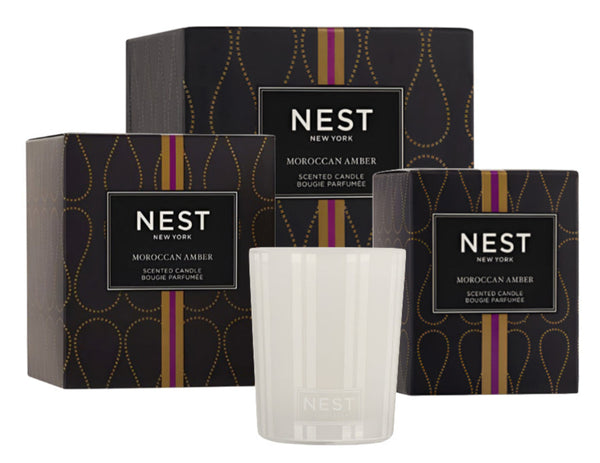 Three Nest NEST Moroccan Amber Candle Collection boxes, infused with bergamot and patchouli, stand elegantly behind a glass candle. Their decorative packaging offers a fragrant journey to Morocco's vibrant markets.