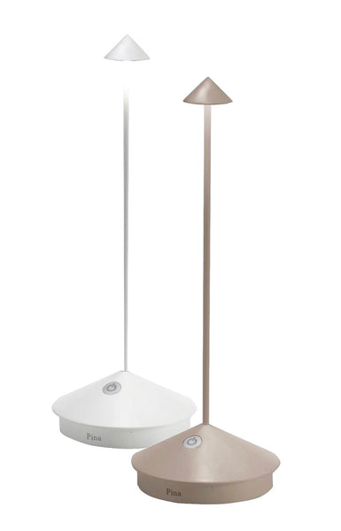 Two modern, touch-dimmable Zafferano Pina Cordless Lamps, one in white and one in beige, with triangular bases and sleek conical shades over LED lights. Each rechargeable lamp from Zafferano America features a touch-sensitive button on the base.