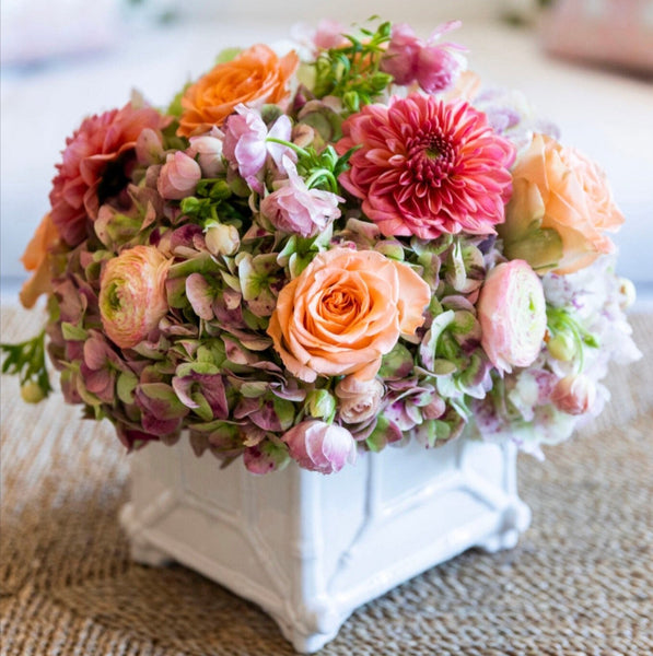 The Live Fall Floral Arrangement by Hive Floral Studio features a stunning mix of pink dahlias, peach roses, and assorted floral accents beautifully presented in a ceramic cachepot on a woven surface.