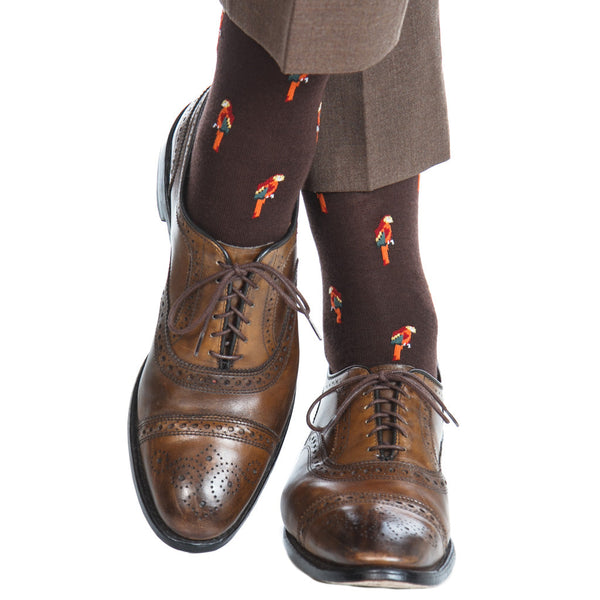 Close-up of a person wearing Dapper Classics brown dress shoes and brown pants, complemented by Dapper Classics Parrot Mid Calf Socks in coffee brown with vibrant orange and red birds, made from breathable material.