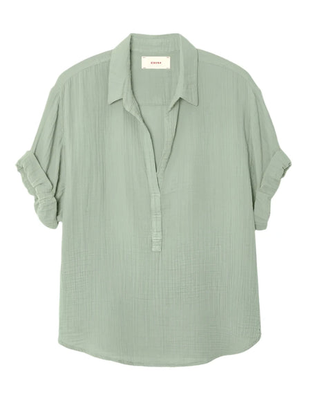 The Xirena Cruz Top by Xirena is a light green, long-sleeve blouse featuring a crinkled texture, a collar, and a partial button-up front made from double-faced cotton gauze. The sleeves are rolled up.