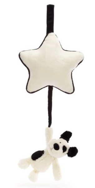 A Jellycat Bashful Black & Cream Puppy Musical Pull with soft cream fur hangs by one arm from a ribbon attached to a soft star-shaped cushion, perfect for comforting a newborn.