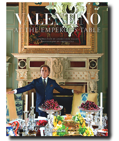 A man in a suit is seated at a lavish table with ornate decor and fruit, as featured on the cover of "Valentino: At the Emperor's Table" by Assouline.