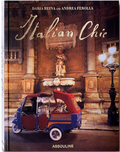 Cover of the product "Italian Chic" by the brand Assouline. The image captures timeless Italian style with a three-wheeled vehicle parked in front of an ornate, lit-up building with a statue. Published by Assouline.