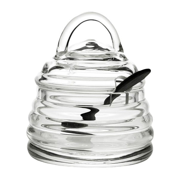 A William Yeoward Crystal Honeybee Honey Jar and Spoon with a spoon in it. The honey jar is reminiscent of a beehive, complete with its hexagonal shape.