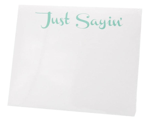 A white notepad from Black Ink's "Black Ink Large Notepad, Just Sayin'" collection rests beautifully in a sleek Lucite Notepad Holder, adorned with the phrase "Just Sayin'" in elegant green cursive at the top, crafted from high-quality paper to capture your thoughts exquisitely.