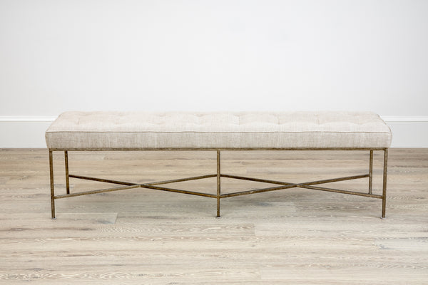 A Jonathan Bench from Oly, featuring a tufted cushion and an antiqued silver finish x-shaped base, sits on a wooden floor against a plain white wall.