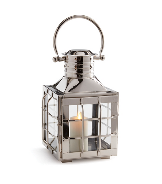 The Nantucket Nickel Outdoor Lantern, Small by Napa Home & Garden features a metallic finish and a handle, housing a lit candle visible through its glass panels and adorned with subtle maritime shapes.