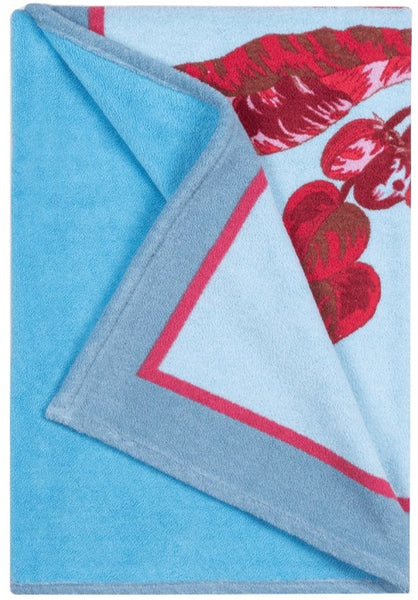 A light blue blanket with a sumptuous cotton terry feel and red floral design on one edge. The blanket, which is partly folded to display both its plain side and the patterned edge, is the Matouk Tallulah Palm Beach Towel in Paradise Pink by Matouk.