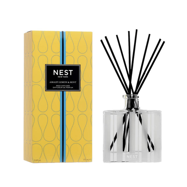 A NEST Amalfi Lemon & Mint Diffuser, by Nest, is shown. The clear glass bottle contains black reeds and sits beside its yellow box packaging adorned with a black label and blue stripe, evoking the fresh, zesty lemon essence of the Amalfi Coast.