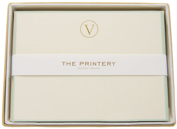 The image displays a box of the Printery Note Card Box Set, showcasing crème-colored stationery with a gold-engraved "V" monogram and "THE PRINTERY, Oyster Bay-New York" printed on the band holding the items together. The ecru initial "V" note card with an aqua border adds an elegant touch to this refined set from Printery.