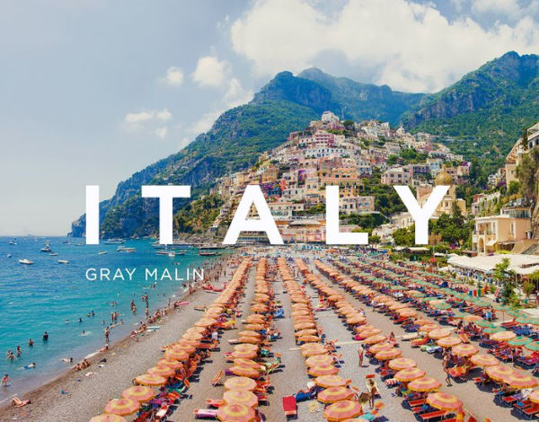 Colorful beach umbrellas line the shore with a picturesque scene from Gray Malin: Italy climbing the hillside in the background.