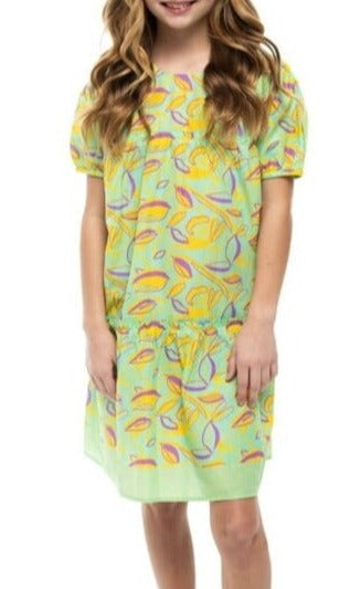 A girl with long, wavy hair stands against a white background, wearing the Briton Court Girls' Meadows Kit Dress from Briton Court, a lightweight cotton green dress adorned with a colorful Meadows print.