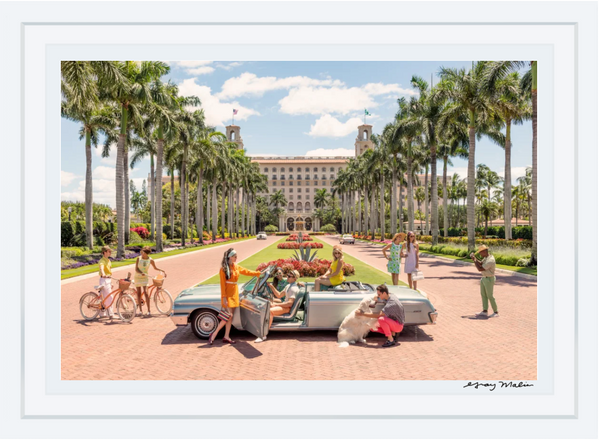 A group of people dressed in colorful clothing gather around a classic car at the entrance to THE BREAKERS PALM BEACH, a grand building with palm trees lining the driveway, evoking the vibrant scenes captured in Gray Malin's *The Breakers Palm Beach* series, available as a framed print measuring 15.5” x 21”.